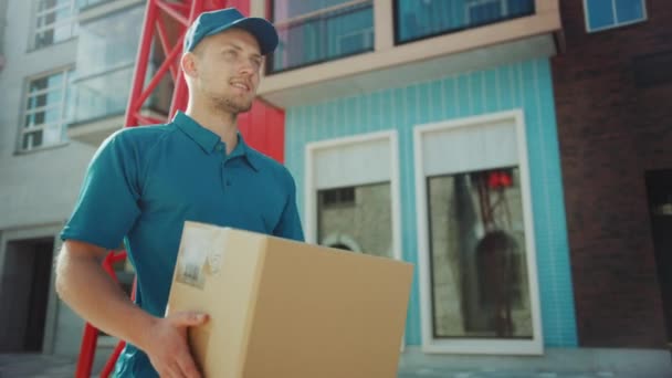 Jóképű Delivery Man tart kartondoboz csomag séta a modern stílusos üzleti negyed. Courier útközben a postai küldemények kézbesítése a kliensre. Alacsony látószögű lassú mozgás — Stock videók