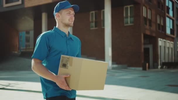 Homem de entrega bonito detém pacote de caixa de papelão caminha através de moderno elegante distrito de negócios. Correio a caminho de entregar encomendas postais a um cliente. Movimento lento de ângulo baixo — Vídeo de Stock