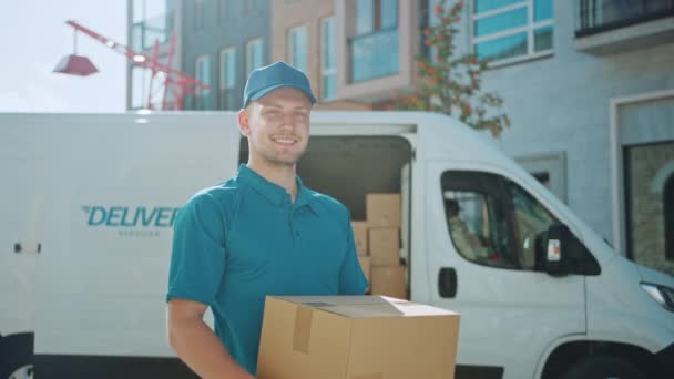 Portrait de bel homme de livraison tient boîte en carton debout dans un quartier d'affaires élégant moderne avec fourgon de livraison en arrière-plan. Coursier souriant en route pour livrer le colis postal au client — Video