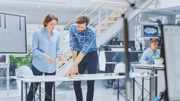 In the Industrial Engineering Facility: Young Female Designer Works with Chief Industrial Engineer, They have Discussion, Analyse and cort Engine Design Technical Drafts that Lying on the Table — 스톡 사진