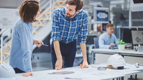 Dans l'installation de génie industriel : Jeune designer travaille avec l'ingénieur industriel en chef, Ils ont des discussions, analyser et corriger la conception du moteur des ébauches techniques qui reposent sur la table — Photo