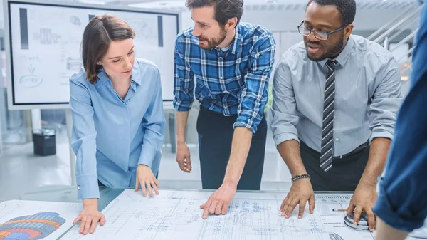 In the Industrial Engineering Facility: Diverse Group of Engineers and Technicians on a Meeting Gather Around Table Unravel Sheets of Engine Design Technical Drafts, Have Discussion, Analyse Drawings