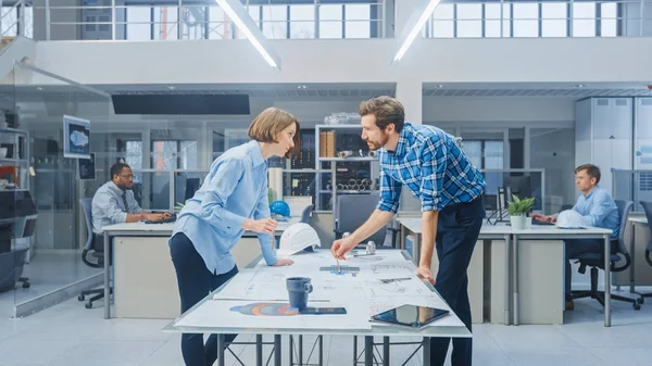 Im Industrial Engineering Facility: Junge Designerin arbeitet mit dem Chefingenieur der Industrie zusammen, sie diskutieren, analysieren und korrigieren technische Entwürfe für die Motorenkonstruktion, die auf dem Tisch liegen — Stockfoto