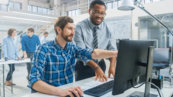 Na Instalação de Engenharia Industrial: Retrato do Bonito Engenheiro Masculino Trabalhando no Computador Desktop, o Engenheiro Chefe se destaca ao lado e explica as particularidades da tarefa e detalhes do projeto. — Fotografia de Stock