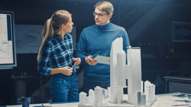 Yaratıcı Mimarlık Bürosu 'nda İki Profesyonel Mühendis Şehir Bölgesi Modeli üzerinde çalışıyor. Şehir Planlamacıları İşlevsel İnşaat Modeli üzerinde çalışıyor. Konuşuyorlar ve Dijital Tablet Bilgisayarı kullanıyorlar
