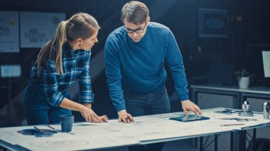 Karanlık Endüstriyel Tasarım Mühendislik Tesisi Erkek ve Kadın Mühendisler Dijital Tablet ve Konferans Masası Kullanarak Bir Planlar Üzerinde Konuşup Çalışıyor. Masaüstü Çizimleri ve Motor Bileşenlerinde