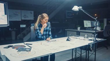 Masasında oturan ve kahvesini içen güzel bayan mühendis. Bir Masanın Üzerinde Yatan Planlar. Karanlık Endüstriyel Tasarım Mühendislik Tesisinde.