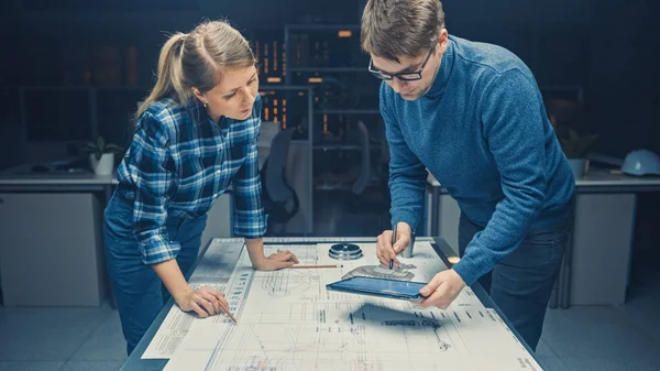 Dans le Dark Industrial Design Engineering Facility Les ingénieurs masculins et féminins discutent et travaillent sur un plan à l'aide d'une table de conférence. Sur les dessins de bureau, les ébauches et les composants du moteur électrique, les pièces — Photo