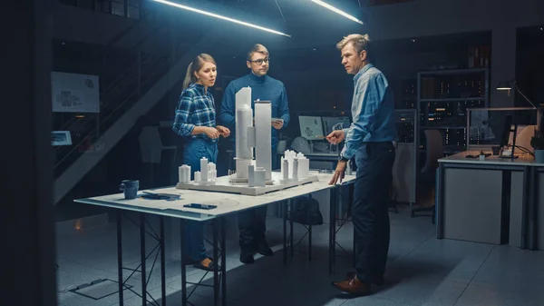 En la Oficina de Arquitectura Creativa, tres ingenieros profesionales trabajan en un modelo de un distrito de la ciudad. Los planificadores urbanos trabajan en un modelo de construcción funcional. Personas que trabajan hasta tarde en la noche — Foto de Stock