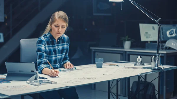 Kvinnlig ingenjör sitter vid skrivbordet Fungerar med ritningar som ligger på ett bord, använder penna, härskare och digital surfplatta. I Dark Industrial Design Engineering Facility — Stockfoto