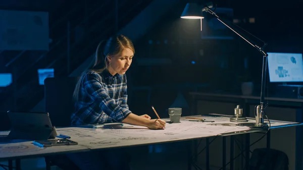 Kvinnlig ingenjör sitter vid skrivbordet Fungerar med ritningar som ligger på ett bord, använder penna, härskare och digital surfplatta. I Dark Industrial Design Engineering Facility — Stockfoto