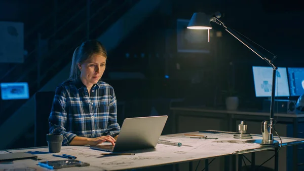 Красива жінка-інженер сидить на столі Працює на ноутбуці комп'ютера. Блакитні відбитки лежать на столі. У темному індустріальному проектному об'єкті. Тепла лампа — стокове фото