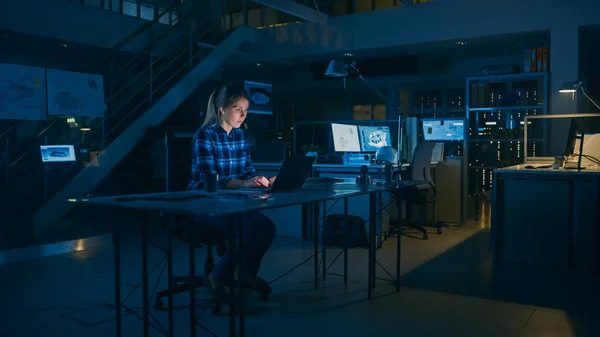 Mooie vrouwelijke ingenieur zit aan haar bureau Werkt op een laptop computer. Blauwdrukken liggend op een tafel. In de Dark Industrial Design Engineering Facility. — Stockfoto