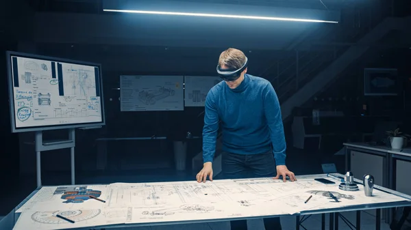 Software Development Engineer Wearing Virtual Reality Headset Gestures and Manipulates Components in Augmented Reality. Engineering Facility Has Desk with Engine and Car Concept Blueprints