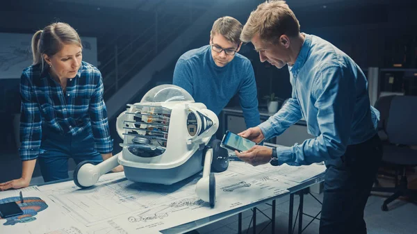 Impianto di ingegneria robotica Tre ingegneri tecnici parlano e lavorano su un prototipo di robot a ruote. Sullo sfondo Centro di Ricerca High Tech con schermi che mostrano disegno industriale. — Foto Stock