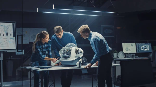 Tarde en la noche en las instalaciones de ingeniería de robótica Tres ingenieros técnicos hablan y trabajan en un prototipo de robot con ruedas. En el centro de investigación de alta tecnología de fondo con pantallas que muestran diseño industrial — Foto de Stock