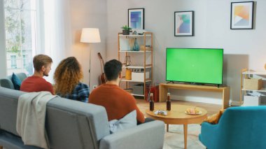 Evde Kanepede oturan üç arkadaş, Atıştırmalık ve içecek içerken yeşil krom ekran televizyon izliyorlar. Gençler Evlerinde Eğleniyor.