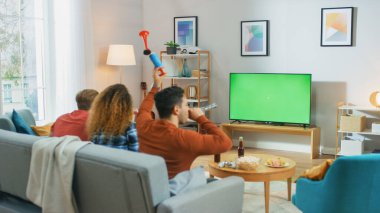 Oturma odasında bir koltukta oturan üç spor fanatiği Yeşil Krom Anahtar Ekran Maçı 'nı izliyor, boynuzlarını kullanıyor, takımları için eğleniyor ve tezahürat yapıyor..