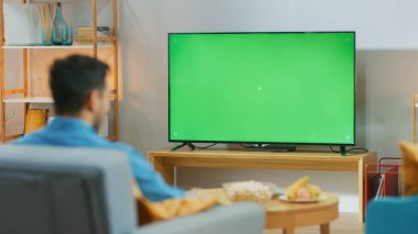 Oturma odasında oturan mutlu adam yeşil krom anahtar ekranını izliyor, işten sonra dinleniyor. Konforlu bir odada Spor Maçı, Haberler, TV Şovu veya Film İzleyen Adam.