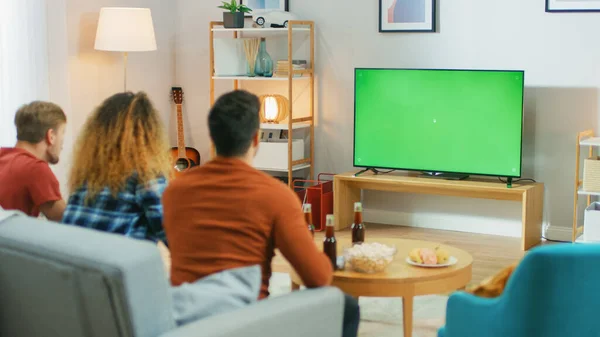 Tres amigos sentados en un sofá en casa, ven el televisor con pantalla clave de croma verde mientras comen aperitivos y beben bebidas. Jóvenes divirtiéndose en casa . —  Fotos de Stock