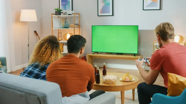En la sala de estar, tres amigos juegan competitivamente videojuegos en la pantalla clave de croma verde, utilizando controladores. Adolescentes divirtiéndose en casa . —  Fotos de Stock