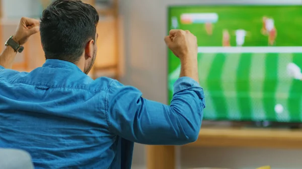 На домашньому спортивному шоу Fan Watches Important Soccer Match on Tv, Cheering For His Team, Celebrates Doing Yes Gesture after the Goal Brings Victory to His Team. Прикольна вітальня. — стокове фото