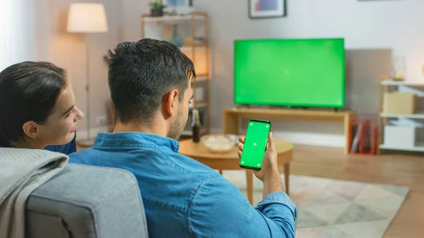 Coppia felice seduta a casa in salotto Guardando verde cromo chiave schermo Televisione, Rilassante su un divano. Guy utilizza anche lo smartphone verde finto schermo . — Foto Stock