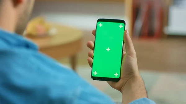 Uomo a casa seduto su un divano tiene verde cromo chiave schermo Touchscreen Smartphone. Naviga attraverso i social network, legge notizie o utilizza App . — Foto Stock