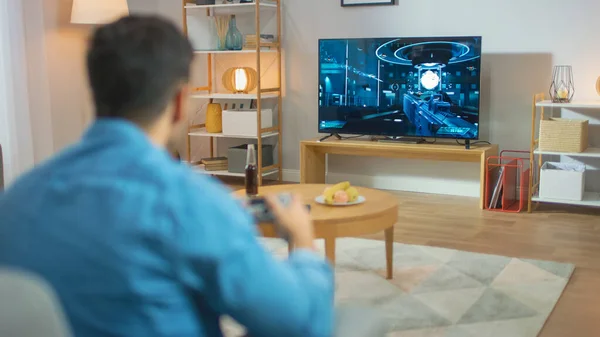 Nel salotto uomo seduto su un divano Holds Controller Giocare in un videogioco di console, 3D azione Shooter Gameplay mostrato sullo schermo della TV . — Foto Stock