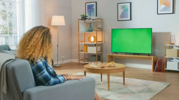 Chica sentada en casa sentada en un sofá, viendo la pantalla clave de croma verde, relajante. Hombre en una habitación acogedora viendo partidos deportivos, noticias, programa de televisión o una película . — Foto de Stock