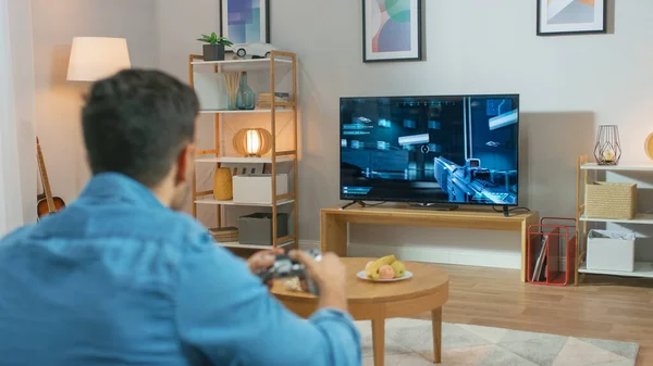 En el hombre de la sala de estar sentado en un sofá sostiene al controlador jugando en un videojuego de consola, la jugabilidad del tirador de acción 3D se muestra en la pantalla de TV . — Foto de Stock