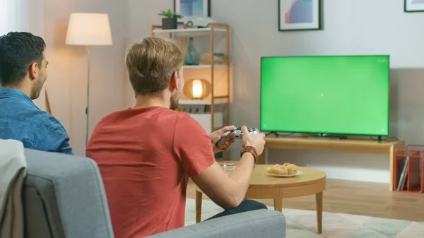En la sala de estar Dos amigos sentados en un sofá que sostiene controladores que juegan a un videojuego competitivo en una pantalla de TV con clave de croma verde . —  Fotos de Stock