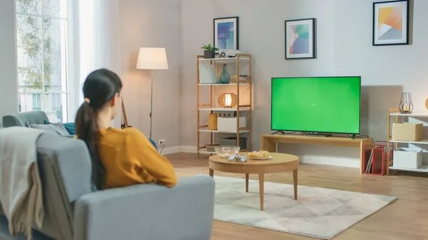 Chica sentada en casa sentada en un sofá, viendo la pantalla clave de croma verde, relajante. Hombre en una habitación acogedora viendo partidos deportivos, Noticias, Sitcom TV Show o una película . — Foto de Stock