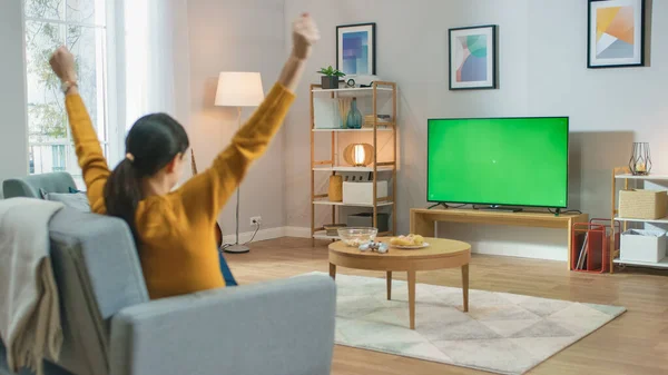 Chica sentada en casa sentada en un sofá, viendo la pantalla clave de croma verde, hace el gesto ganador con los brazos. Hombre en una habitación acogedora viendo partidos deportivos, Noticias, Sitcom TV Show o una película . — Foto de Stock