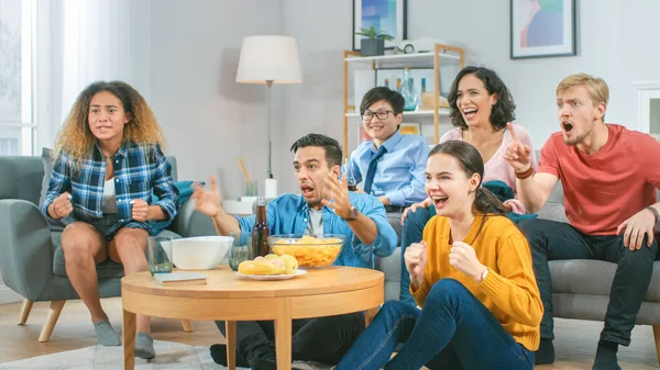 Zu Hause sitzt eine Gruppe von Sportfans auf der Couch und schaut wichtige Spiele im Fernsehen, sie feuern die Mannschaft in einem sehr angespannten Moment an. gemütliches Zimmer mit Snacks und Getränken. — Stockfoto