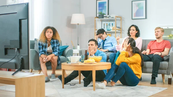 En el grupo diverso de la sala de estar de amigos en el partido del juego de los deportes que miran en la TV, animan y cantan para el equipo, pero se decepcionan después de que el equipo pierde. Acogedora habitación con aperitivos y bebidas — Foto de Stock