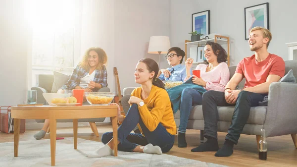 На Home Diverse Group друзі спостерігають за Tv Together, поїдання закусок і пиття. Вони, ймовірно, дивляться "Спортивні ігри, кіно" або "Sitcom Tv Show". Молоді люди розважаються разом. — стокове фото