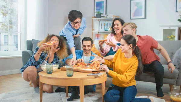 Thuis diverse groep vrienden kijken tv samen, ze delen Gigantic Pizza, eten smakelijke taart stukken. Jongens en meisjes kijken naar komedie sitcom of een film, lachen en plezier hebben samen. — Stockfoto