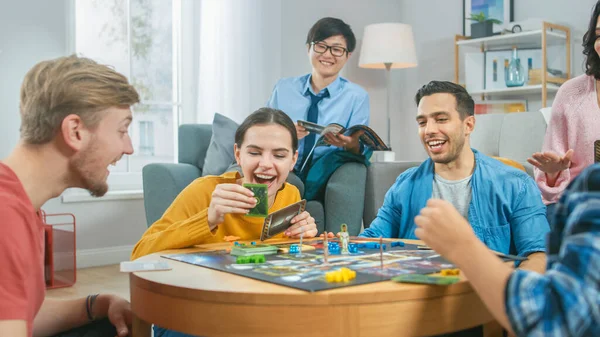 Eine bunte Gruppe von Jungs und Mädchen spielt in einem strategischen Brettspiel mit Karten und Würfeln. Mann würfelt eindrucksvoll. gemütliches Wohnzimmer an einem Tag — Stockfoto