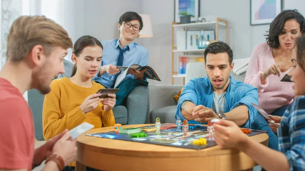 Olika grupp av killar och flickor som spelar i ett strategiskt brädspel med kort och tärningar. Mysigt vardagsrum på en dag — Stockfoto