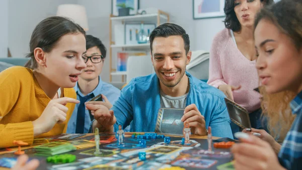 Diverso gruppo di ragazzi e ragazze che giocano in un gioco da tavolo strategico progettato in modo unico con carte e dadi. Amici che si divertono a leggere carte, scherzare, fare mosse e ridere in un accogliente salotto — Foto Stock