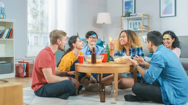 In de woonkamer diverse groep van leuke liefdevolle vrienden spelen "Wie ben ik" spel met plakkerige papieren gehecht aan voorhoofden. Jongens en meisjes proberen te raden wat er op Sticky Notes staat — Stockfoto