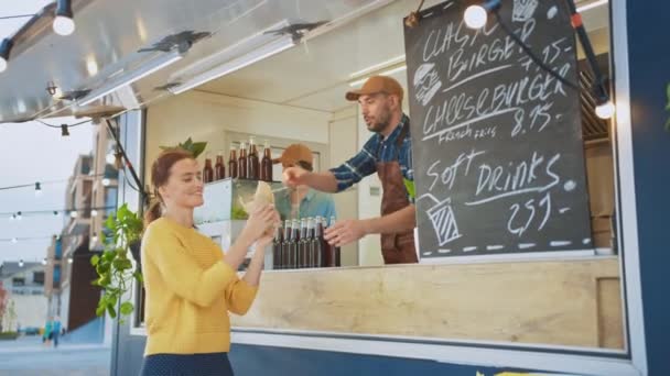 Matbilansatte deler ut en nybakt Gourmetburger til en lykkelig ung kvinne. Unge mann i dress lager en orden. Gatemat - selgende burgere i et moderne nabolag. – stockvideo