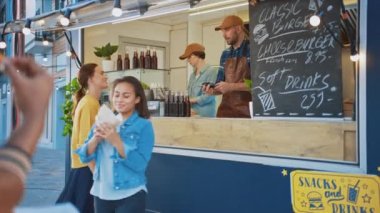 Yemek kamyonu çalışanı genç ve mutlu bir bayana taze yapılmış bir hamburger verdi. Genç bayan yiyecek parasını ödemek için NFC mobil ödemesini kullanıyor. Modern Hip Mahallesi 'nde Sokak Yemekleri Satıyor.