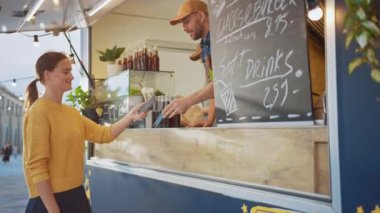 Yemek kamyonu çalışanı Mutlu Genç Bir Kadına Bir Hamburger Verdi. Genç bayan yemek parasını ödemek için akıllı telefonuna NFC Temassız Ödeme yapıyor. Sokak Yemekleri Kamyonu Açık havada Burger Satıyor.