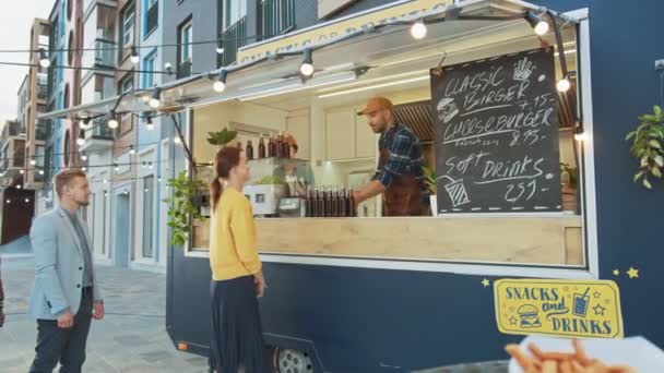 Empleado de Food Truck entrega una hamburguesa gourmet recién hecha a un joven y feliz en cola. Camión de comida callejera vendiendo hamburguesas al aire libre en un barrio moderno de la cadera . — Vídeo de stock