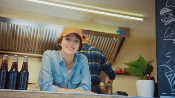 Food Truck Feminino Empregado Sorri e olha para a câmera. Street Food Truck vendendo hambúrgueres em um bairro moderno. Proprietário de negócios de comida de rua de sucesso é feliz no trabalho. Morena usa um boné . — Vídeo de Stock