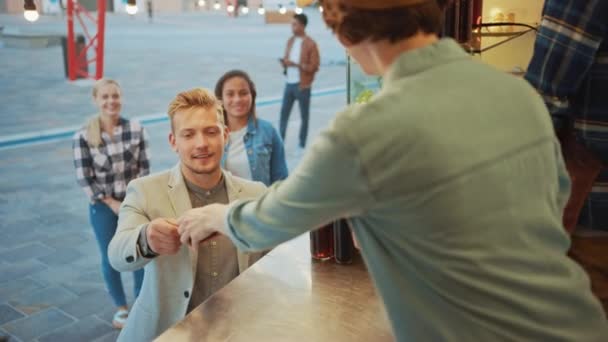 Empleado de camiones de comida entrega una hamburguesa recién hecha a un joven feliz con un traje. Macho está pagando por la comida con tarjeta de crédito sin contacto. Camión de comida callejera vendiendo hamburguesas en un vecindario moderno de la cadera — Vídeo de stock