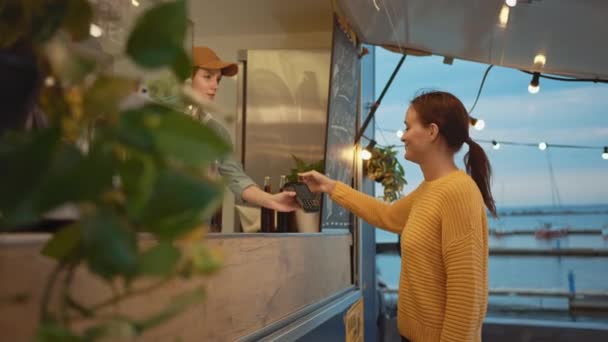 Empleado de camión de alimentos entrega una hamburguesa recién hecha a una joven y feliz hembra. Young Lady está pagando por comida con tarjeta de crédito sin contacto. Camión de comida callejera vendiendo hamburguesas en un vecindario moderno de la cadera — Vídeo de stock