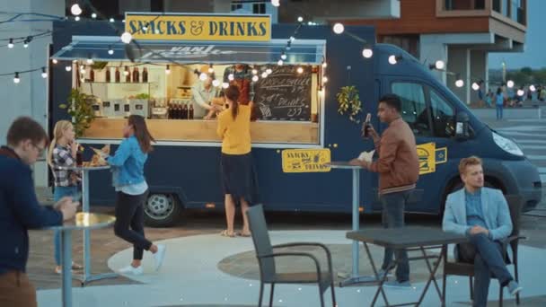 フードトラックの従業員は幸せな若いヒップスターの顧客への新鮮に作られたビーフバーガー、フライおよび冷たい飲み物を手渡す。近代的なクールな近所の商業トラック販売ストリートフード. — ストック動画